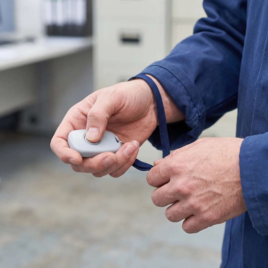 A worker's hand holds a small personal safety alarm with a thumb resting on its button.